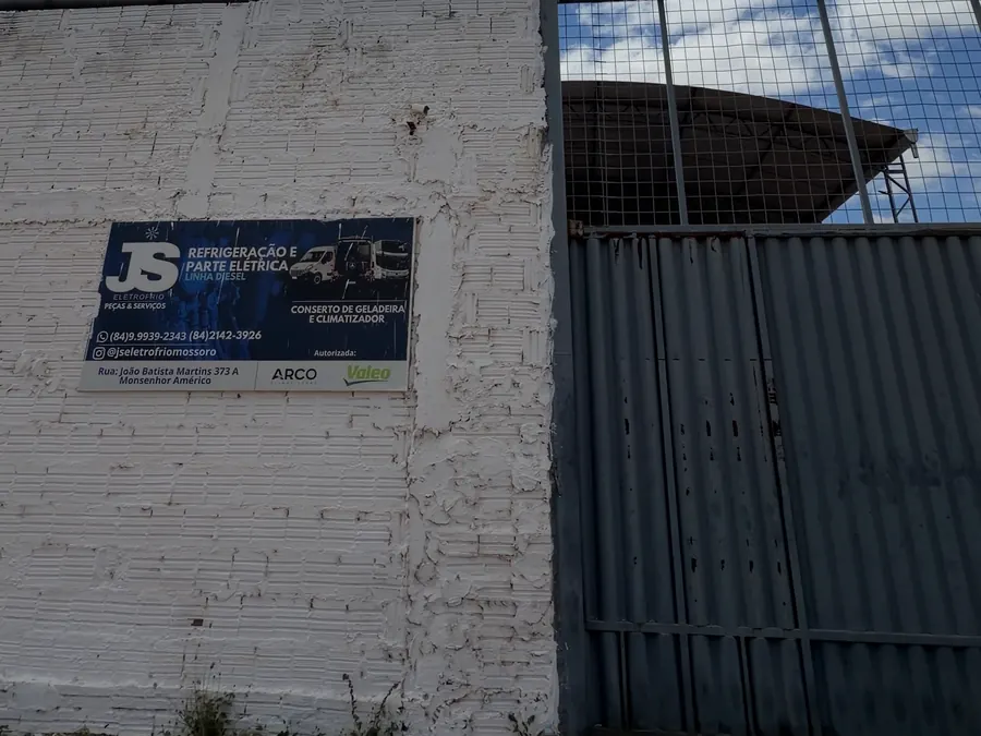 Fachada da JS Eletrofrio: muro branco com a placa da oficina e o portão de entrada, na Rua João Batista Martins, Monsenhor Américo